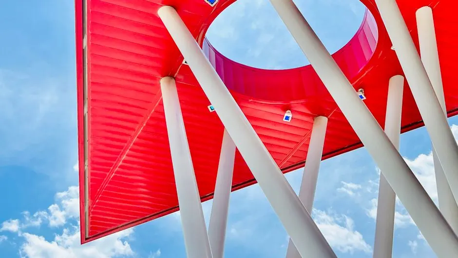 Low angle view of a modern red roof structure with white beams against a blue sky.